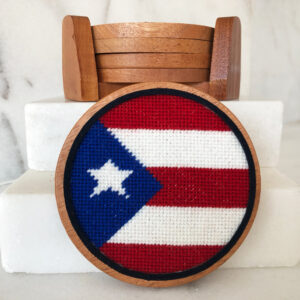 A round wooden coaster with a cross stitch flag of puerto rico.